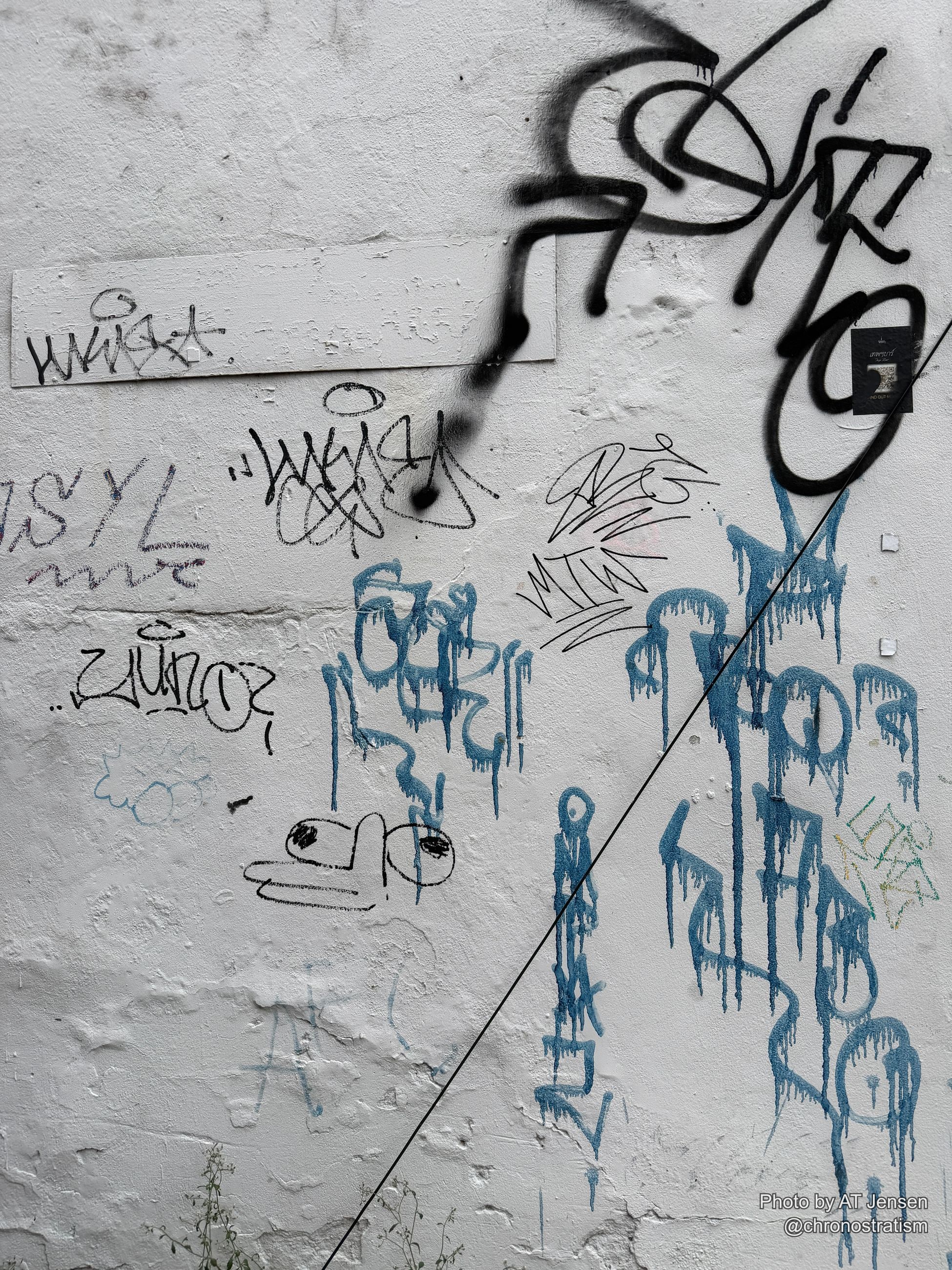 Black spray-painted tags and blue marker scrawl cover a white-painted wall, a wire crossing overhead.