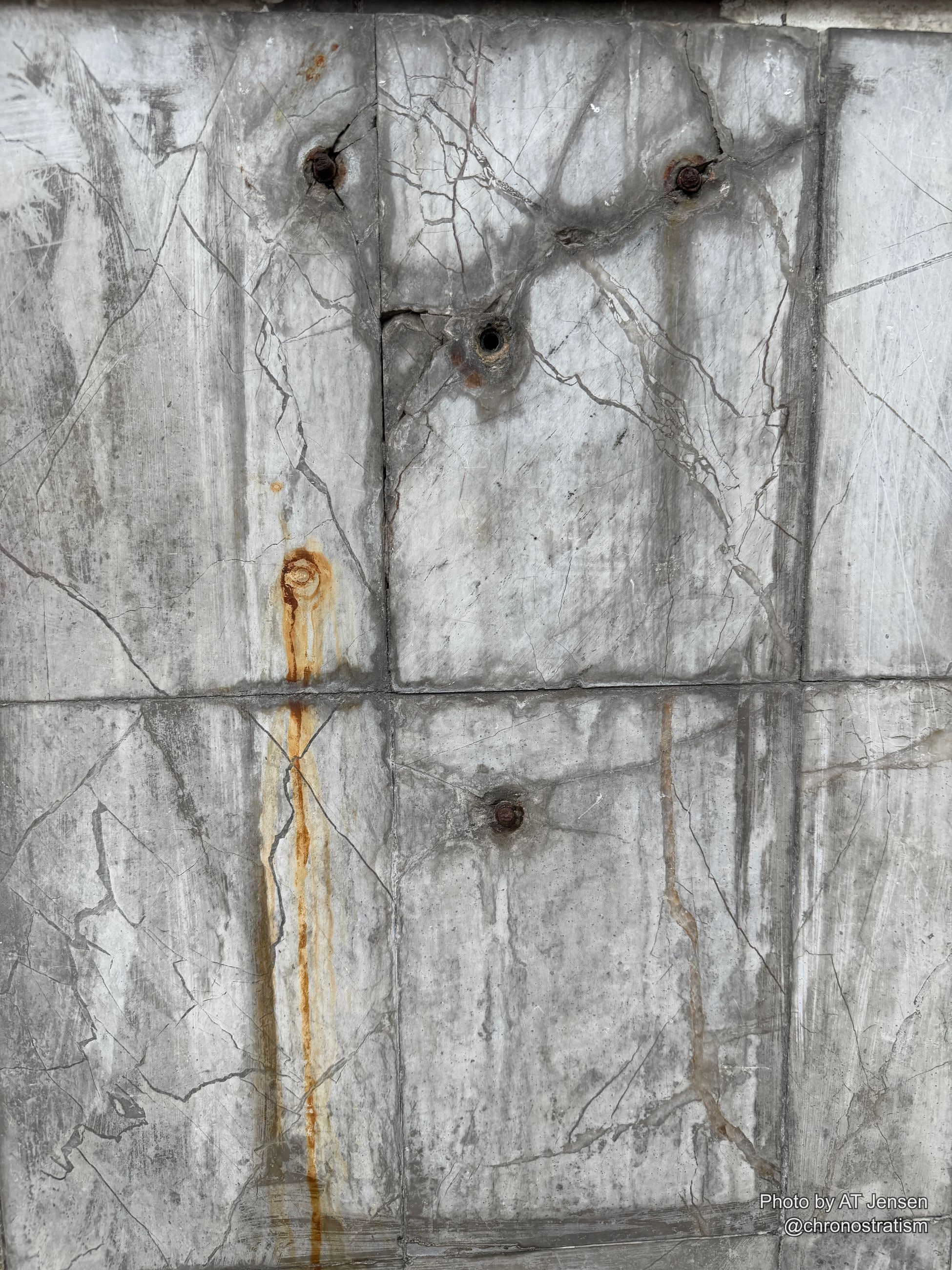 Fine cracks web across polished marble-like tile, rust streaks bleeding from corroded bolt holes.