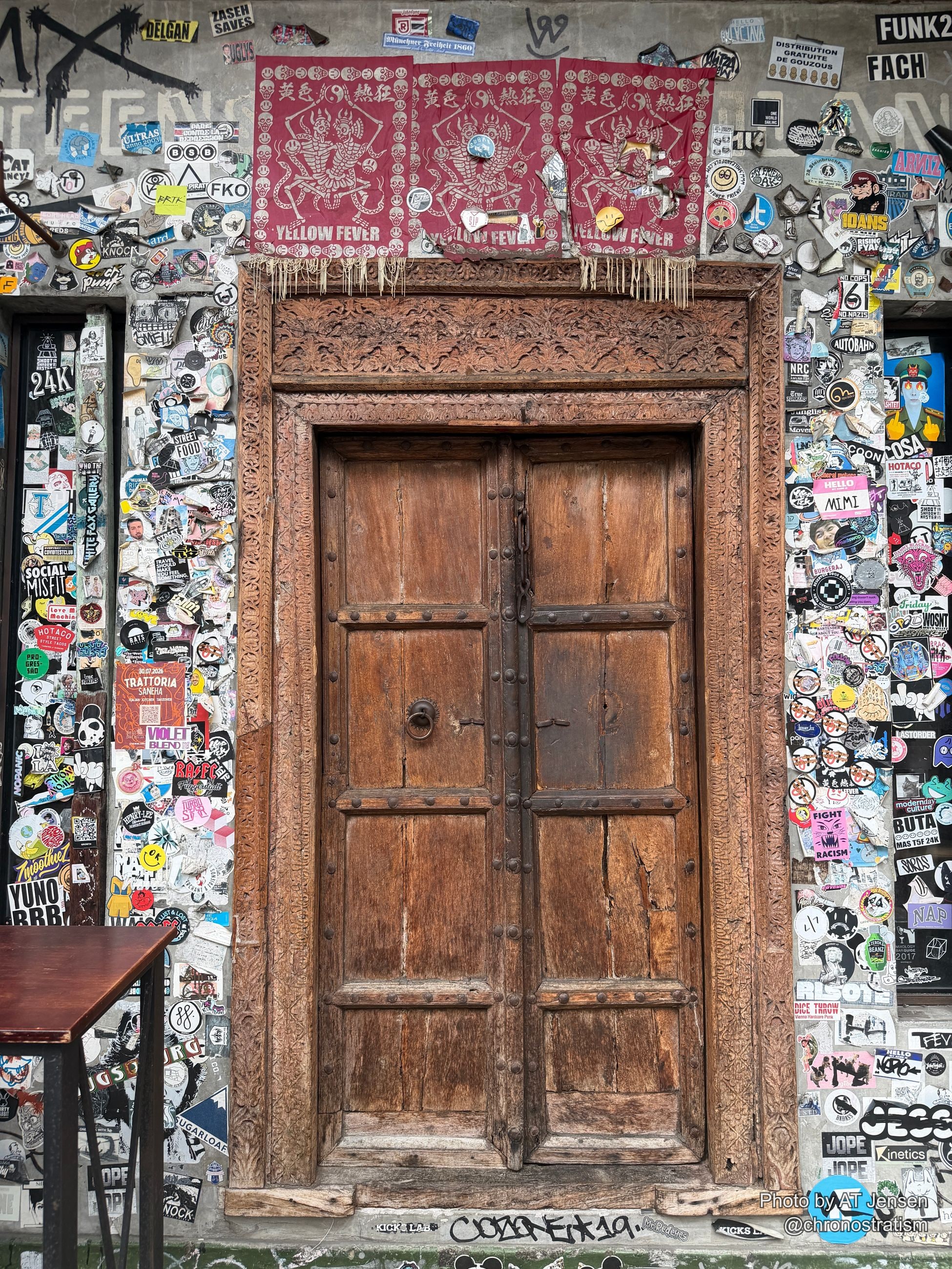 A dense sticker and paste-up collage frames an ornate carved wooden door, a red talisman cloth hanging beside it.