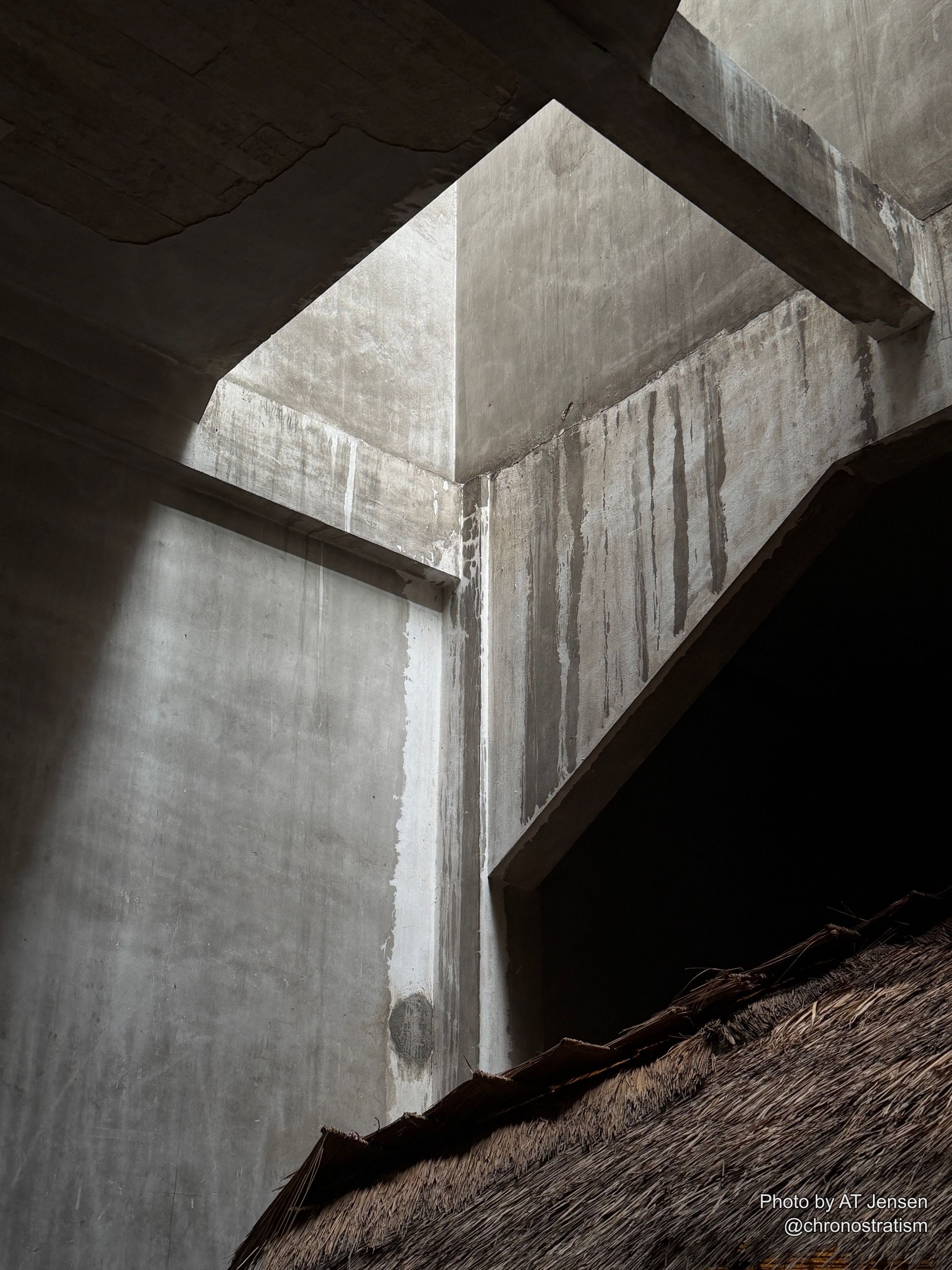Dramatic light rakes across a weathered interior corner where a thatched roof edge meets bare concrete.