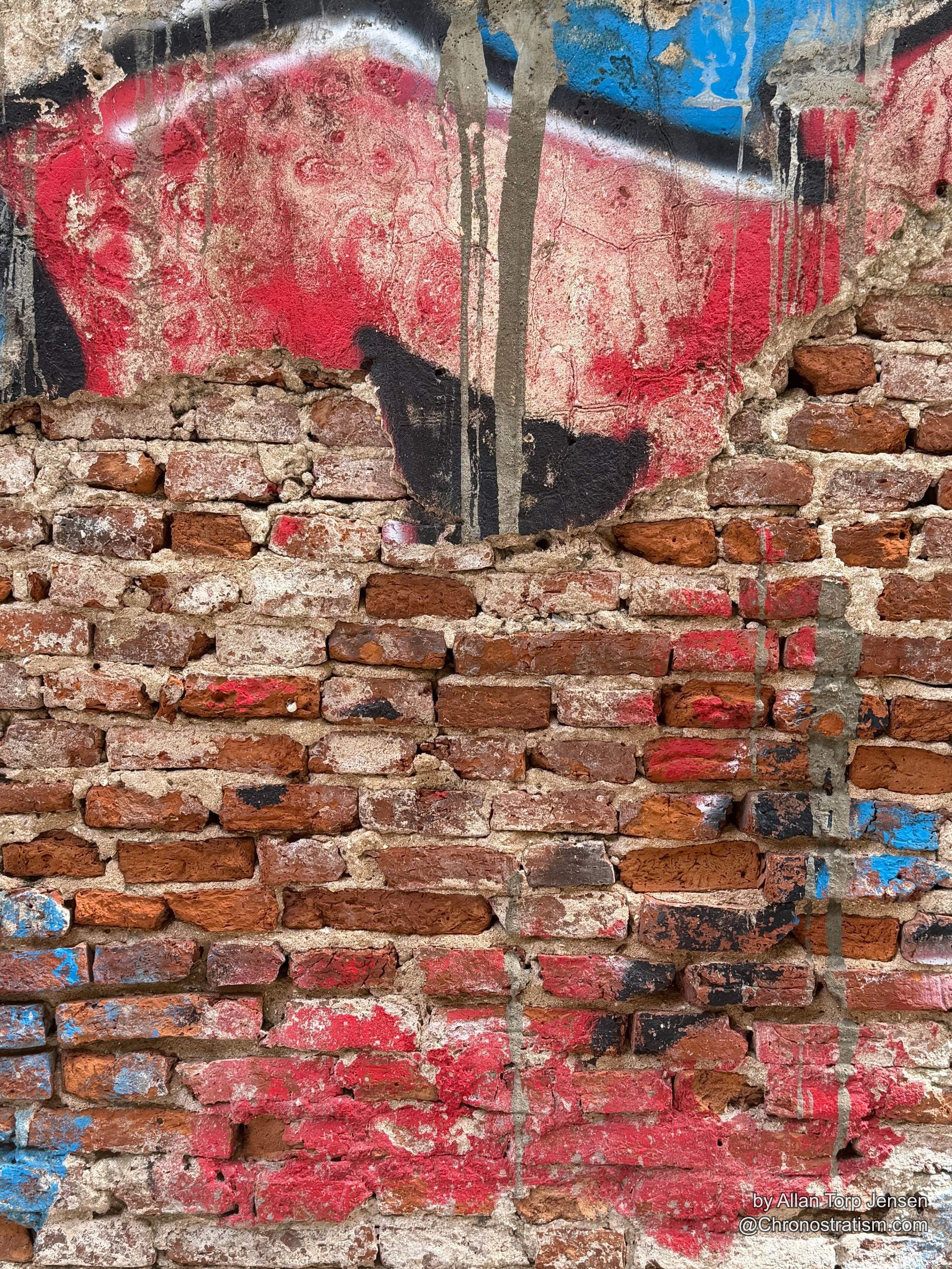 A faded red and blue stencilled figure clings to plaster crumbling toward exposed brick.