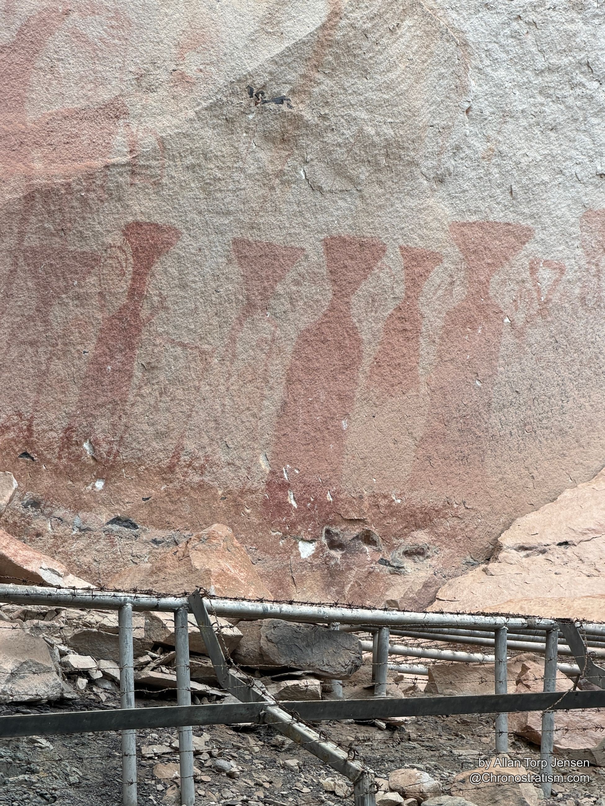 Faded red ochre pictograph symbols mark a weathered stone wall, rusted wire mesh at the edge.