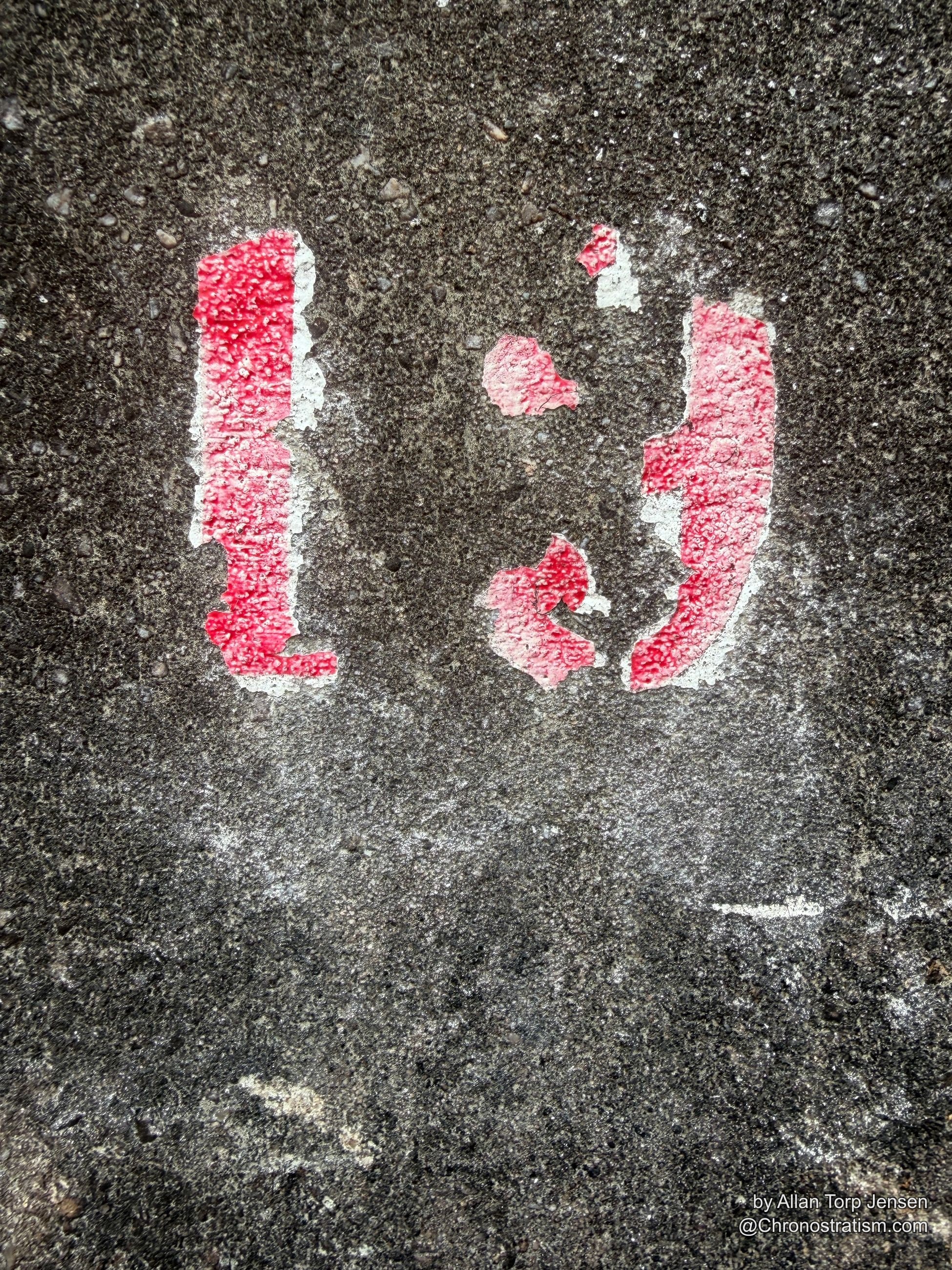 A faded red stencilled mark flakes away on rough dark asphalt, only fragments of the paint surviving.