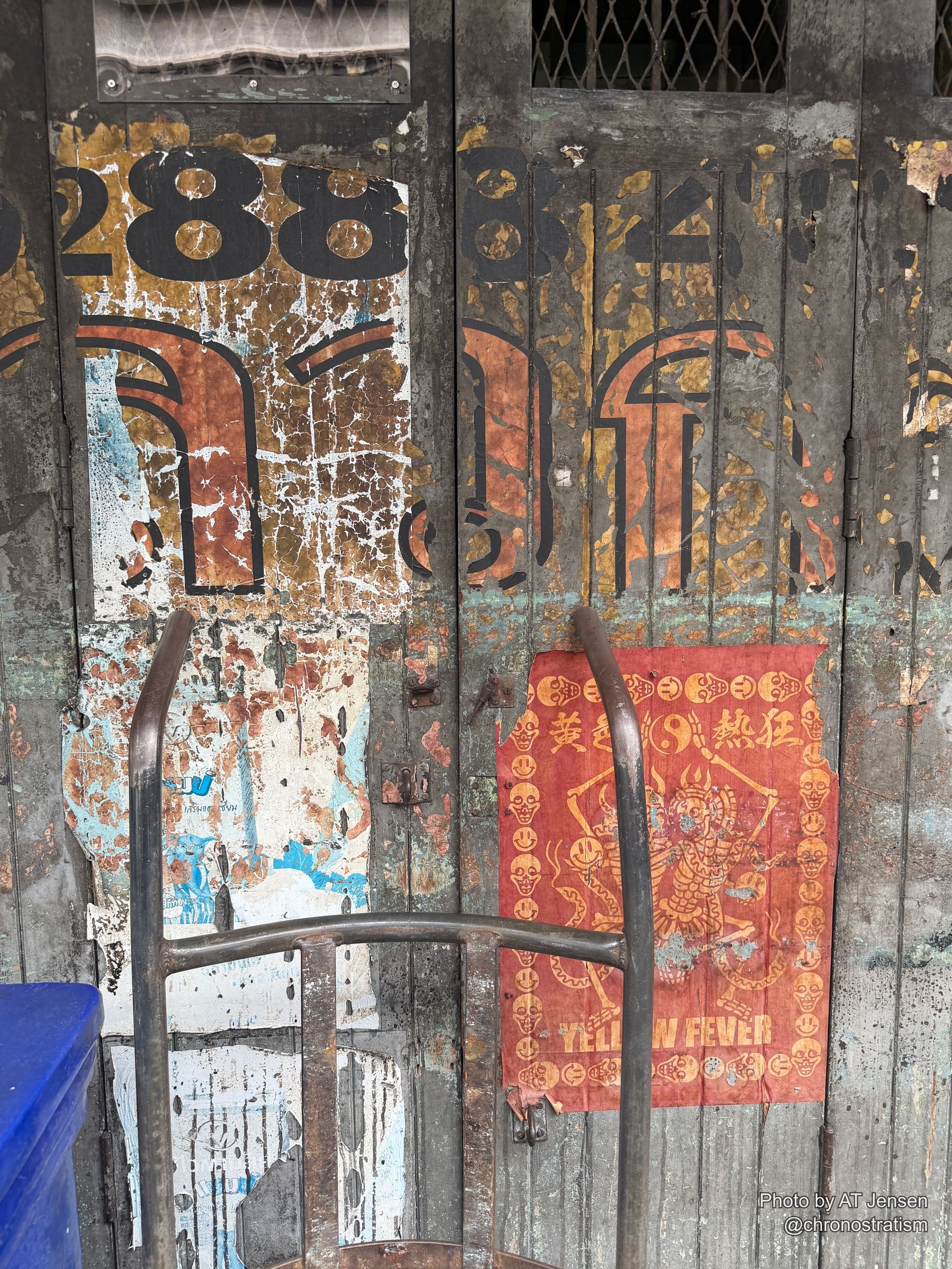 A faded red talisman paper and stencilled numbers cling to a weathered metal shutter.