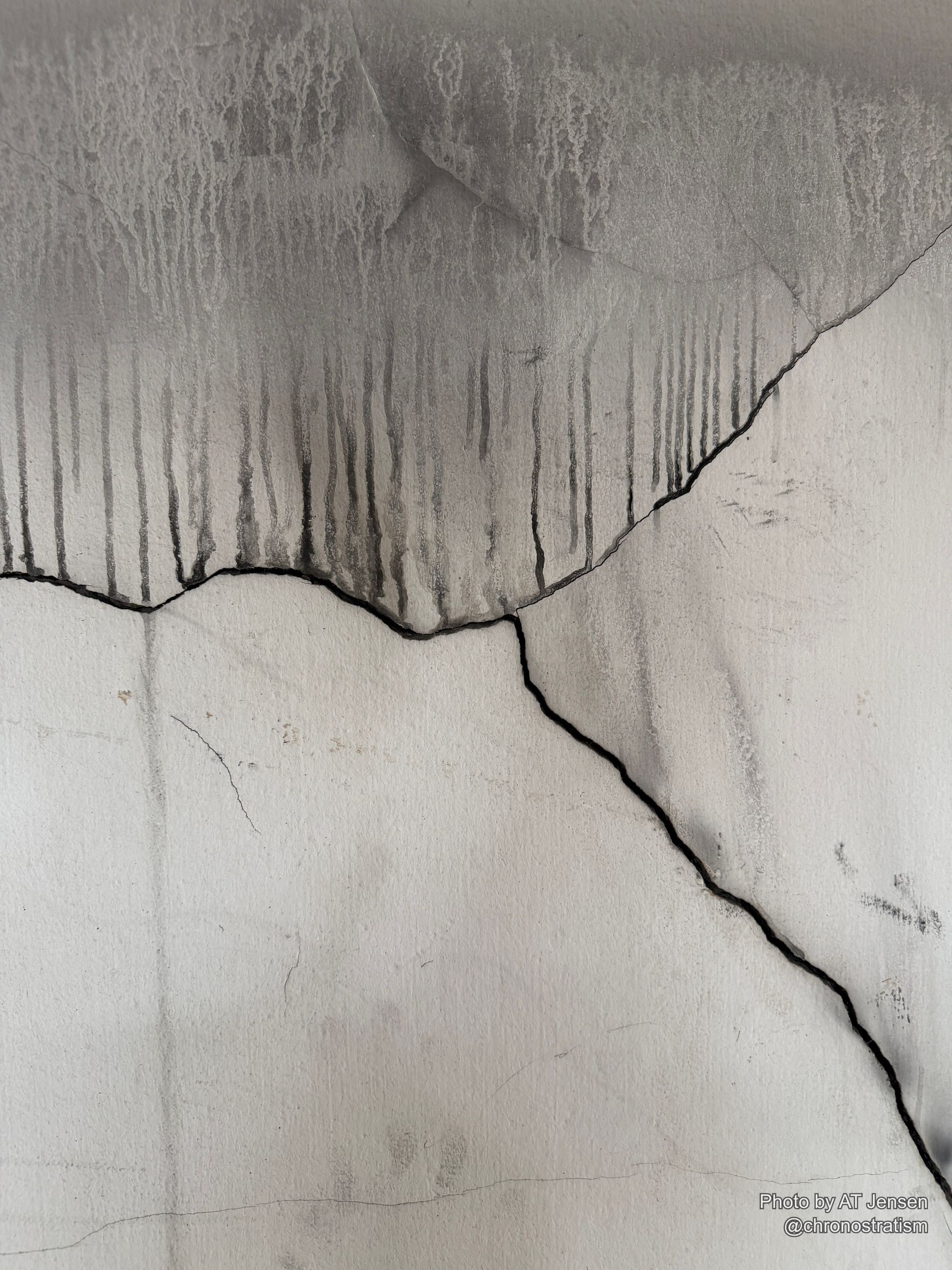 A fine hairline crack cuts diagonally across a two-tone render wall, faint striations running through the paler half.