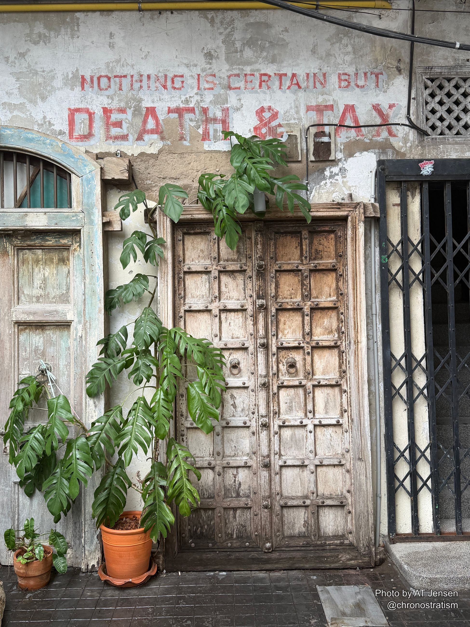 A hand-painted ‘Nothing is certain but death & tax’ sign fades above a pair of old wooden doors framed in greenery.