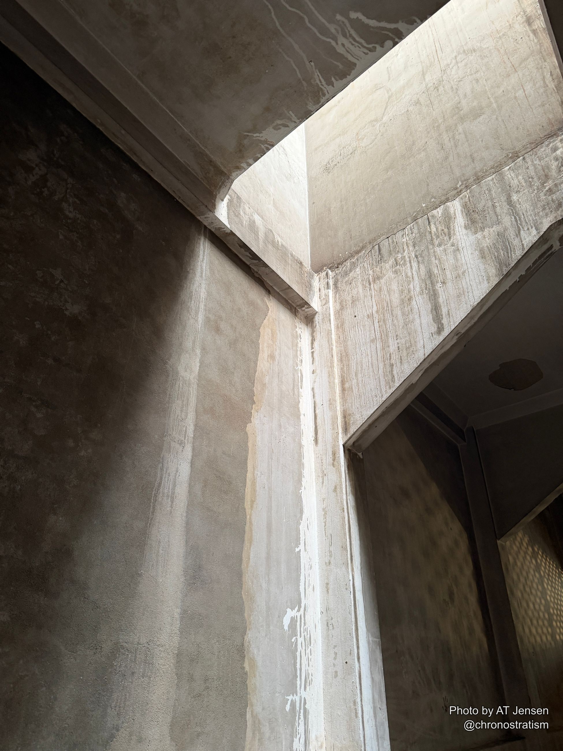 A shadowed interior corner frames a barred grille window against rough concrete walls.