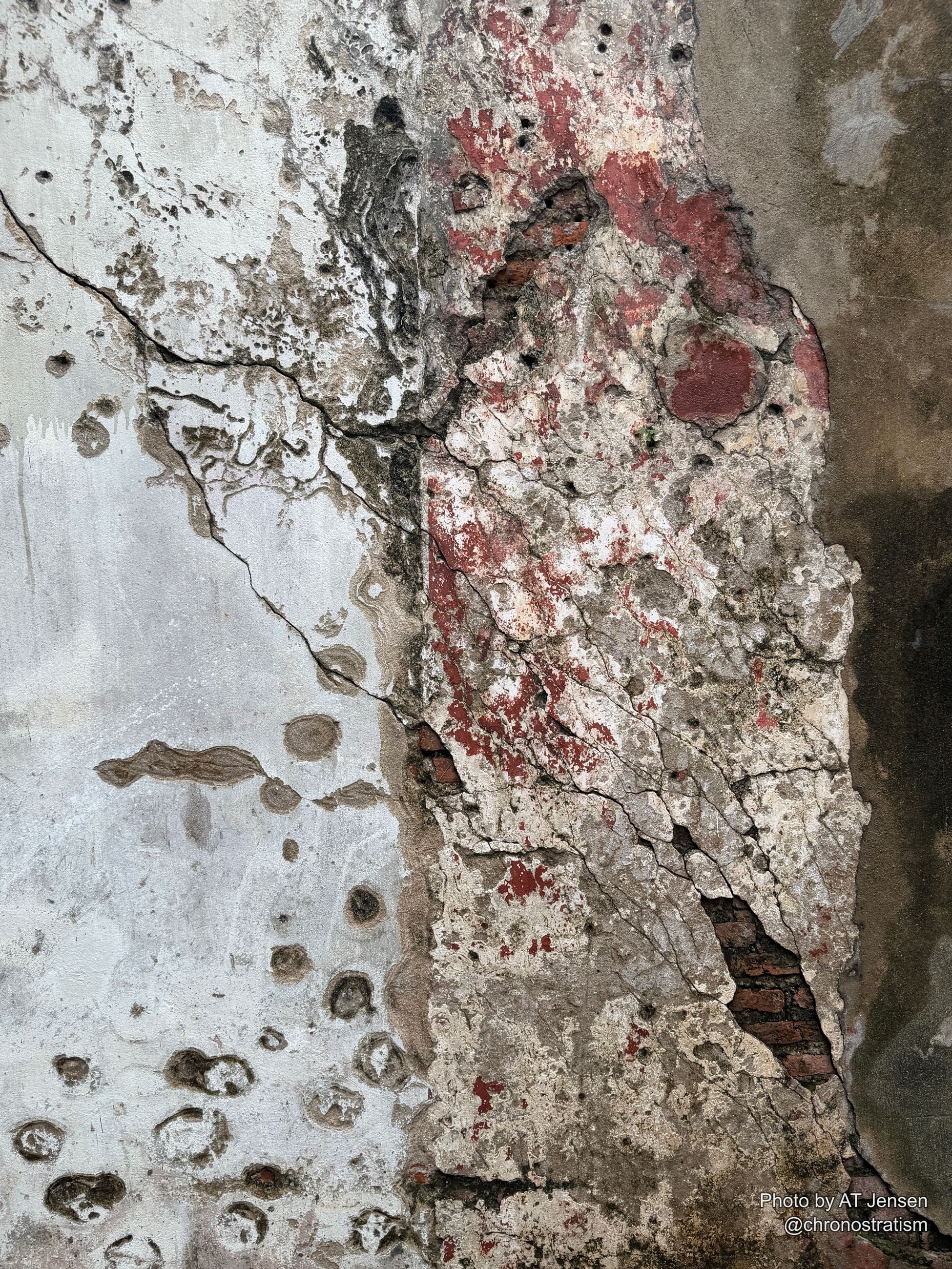 Layered cracked plaster reveals red pigment and brick beneath, giving way to a dense band of pitted, weathered concrete below.