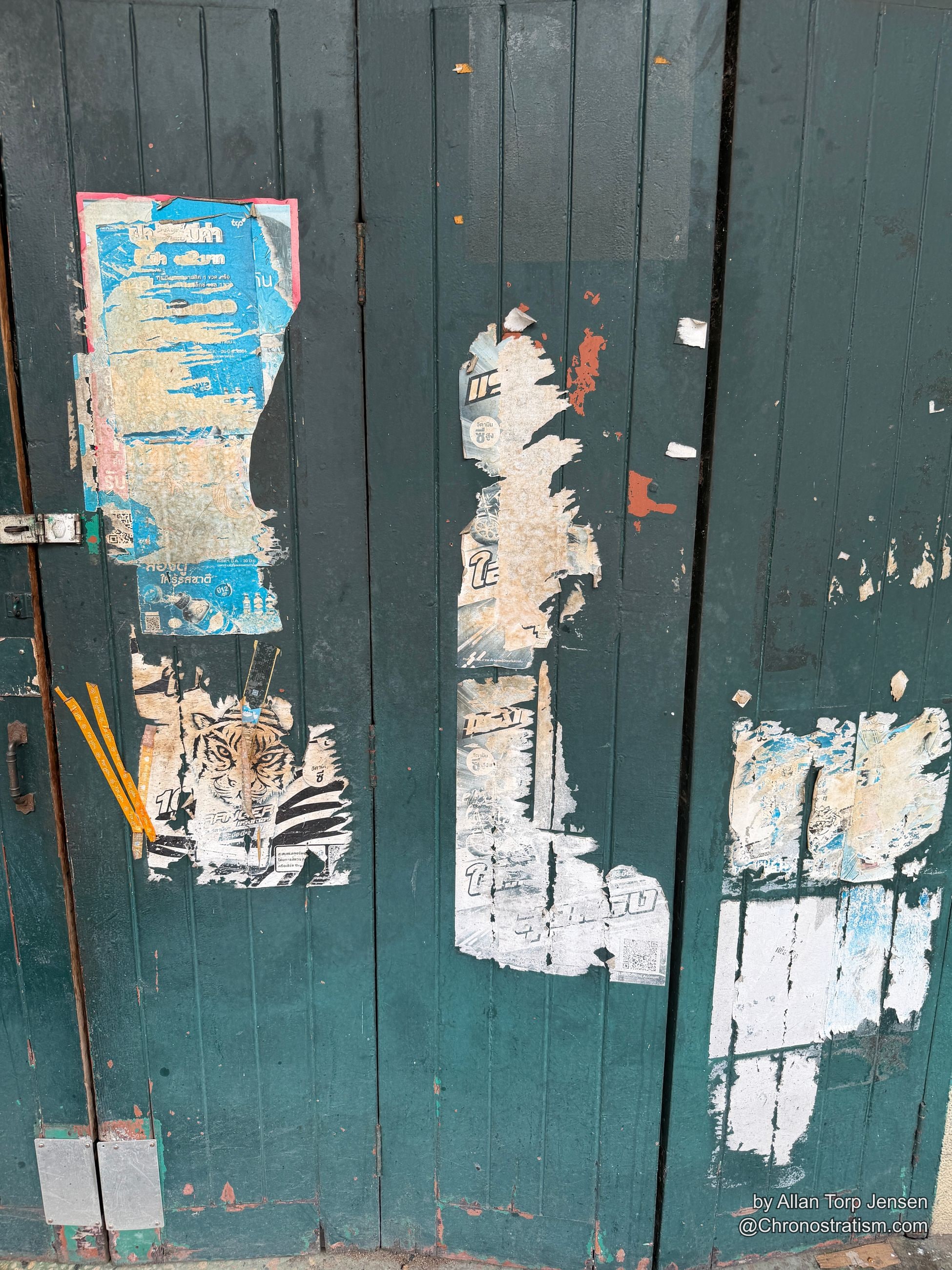 Layers of torn wheatpaste posters cling to a teal wooden door, faded print bleeding through.