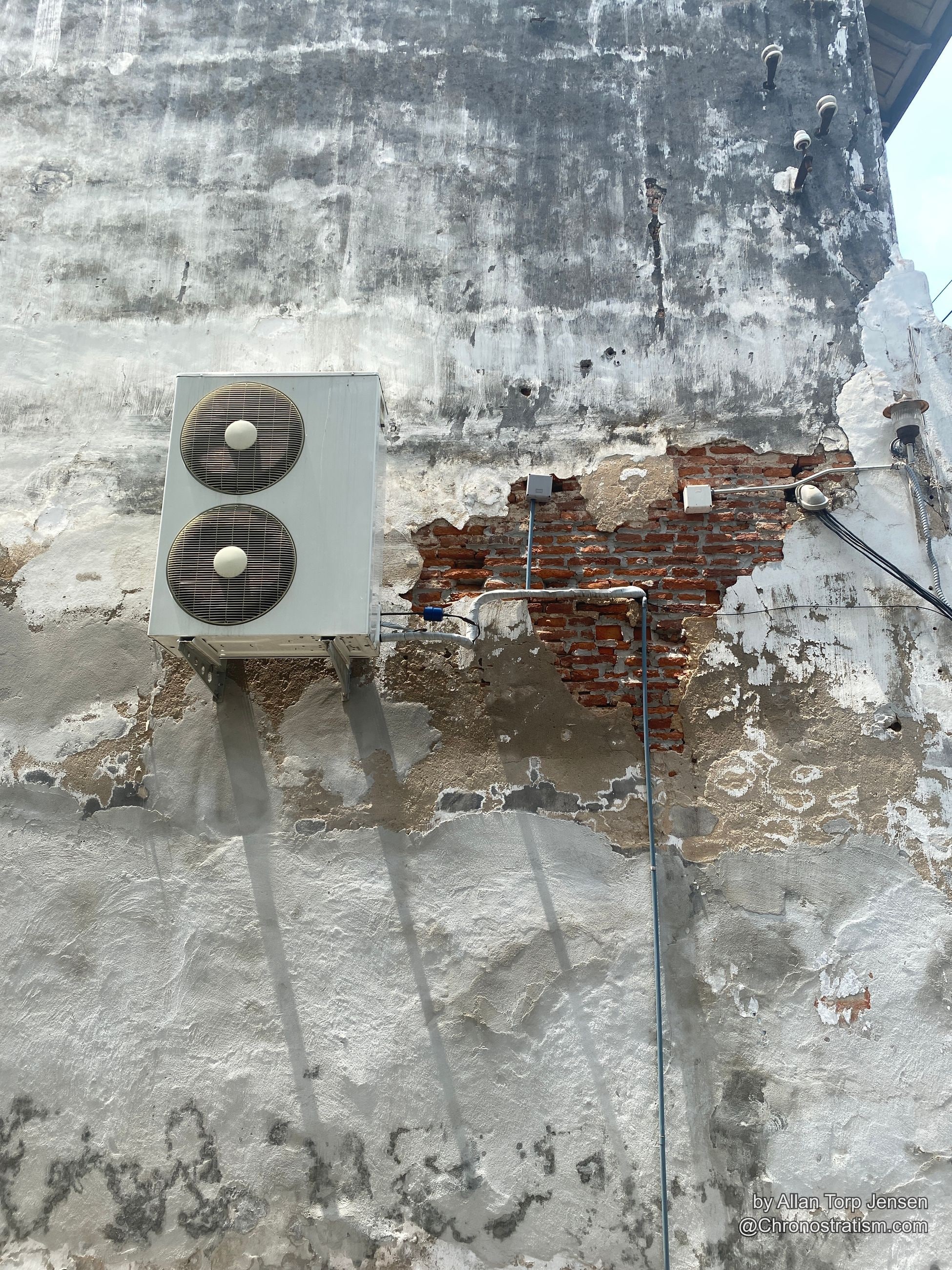 A patch of red brick exposed beneath crumbling white render, conduit pipes crossing the scar.