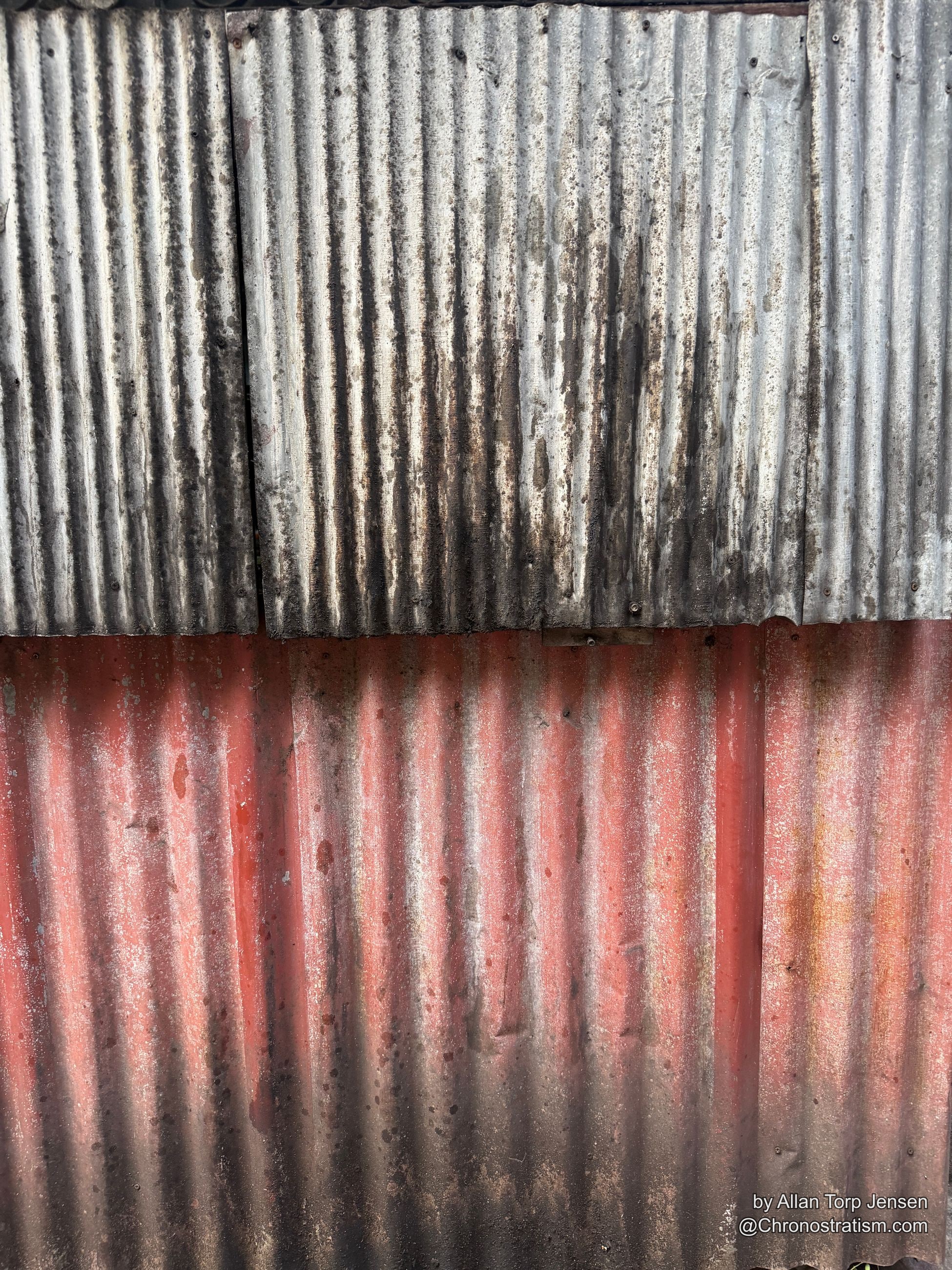 Rust bleeds through red paint on one half of a corrugated metal fence, soot streaking the other.