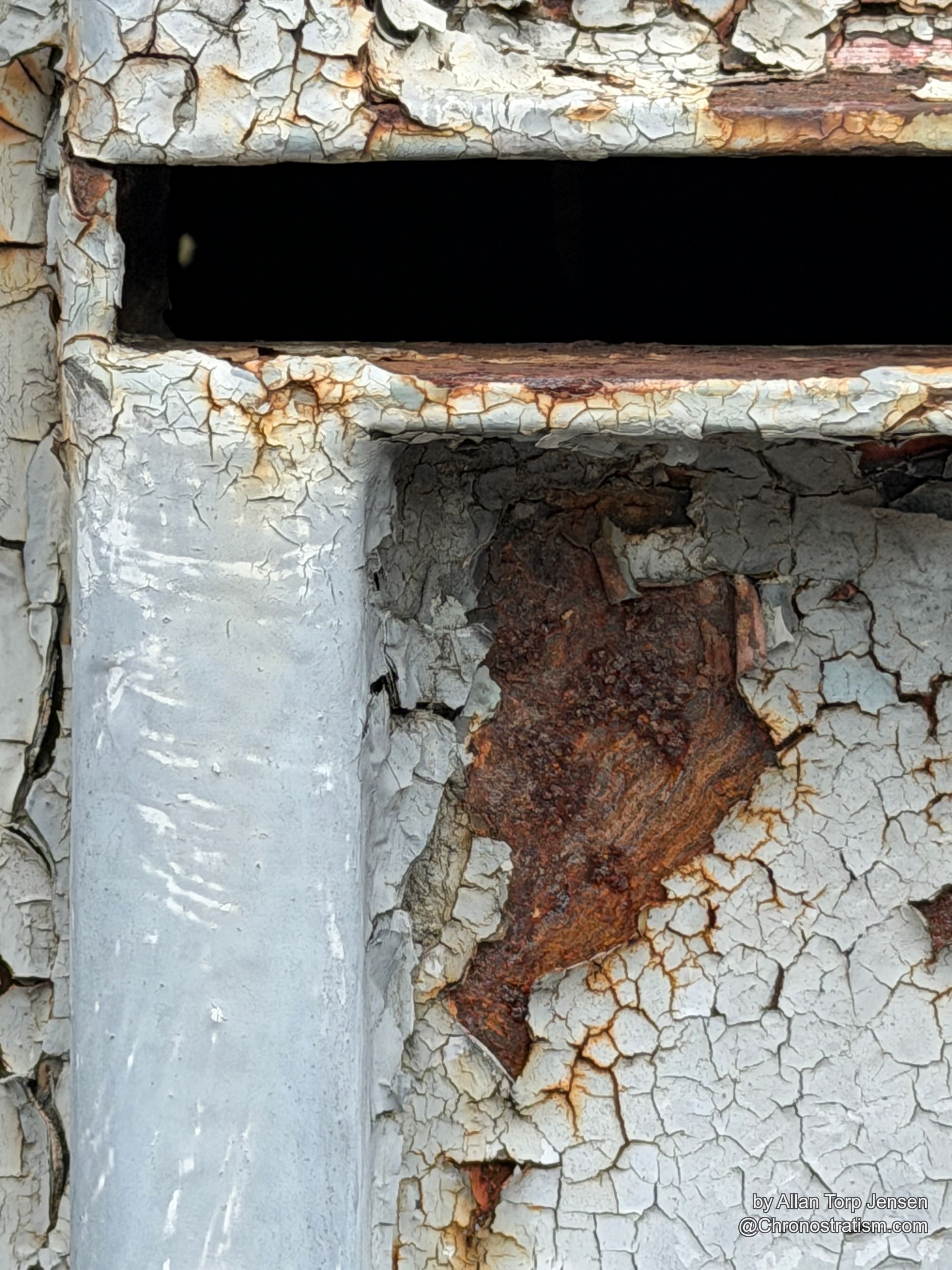 Rust blooms through a peeling white door where paint has curled back near a dark slot opening.