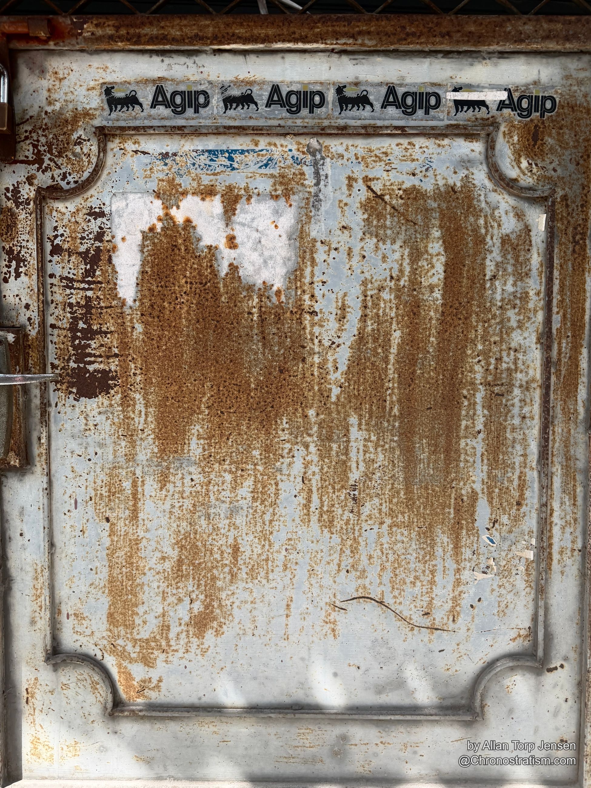 Rust streaks bleed down a metal door plastered with repeated Agip oil-brand stickers.