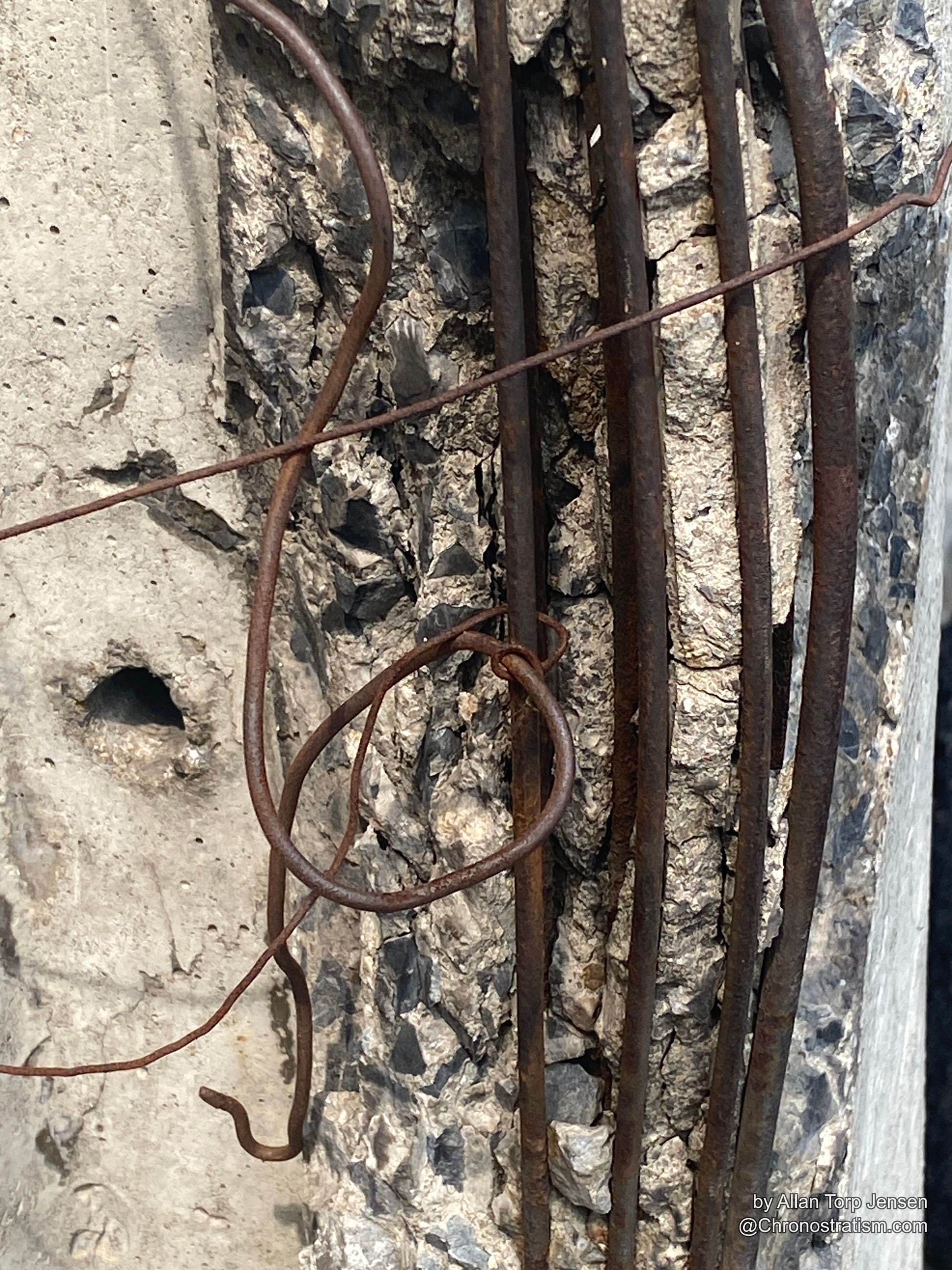 Rusted rebar loops through a spalling concrete edge, the structure fracturing from within.