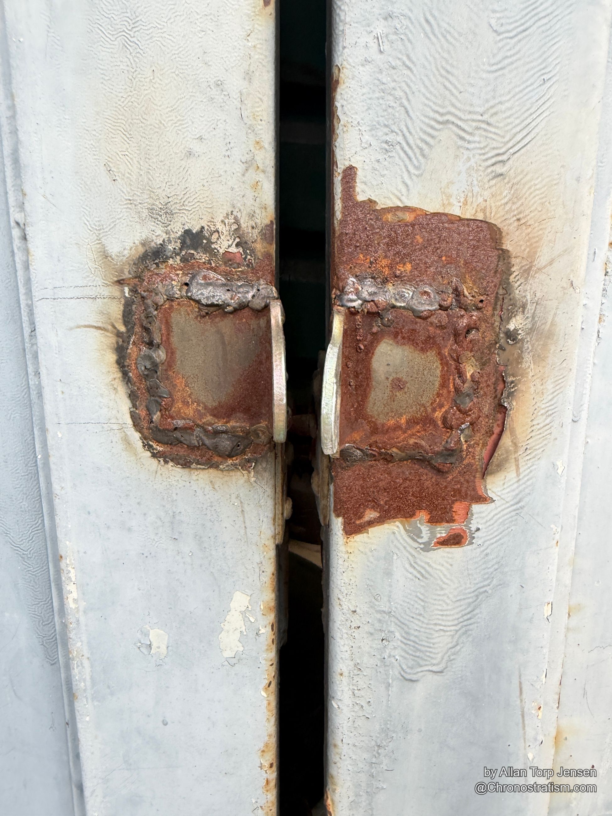 A rusted weld patch scars a white-painted steel beam, twin square repairs bolted in place.