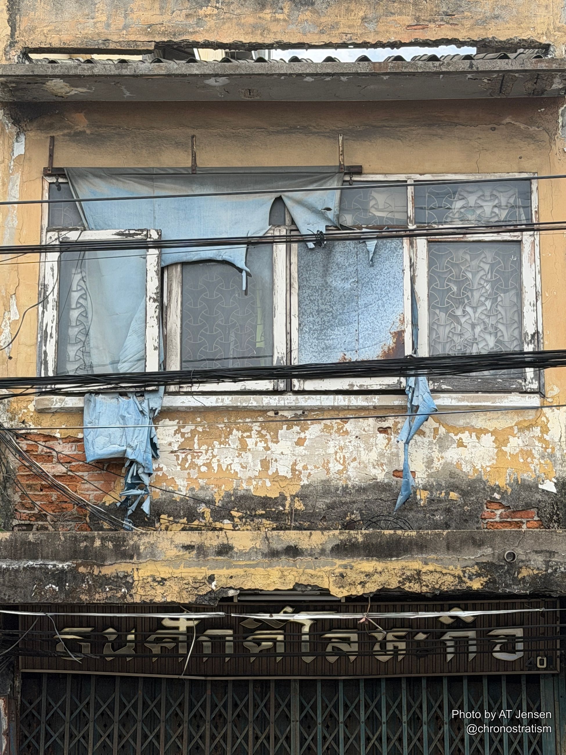 Tangled wires and torn plastic tarps cover a shophouse window, peeling paint and grime streaking the wall around it.