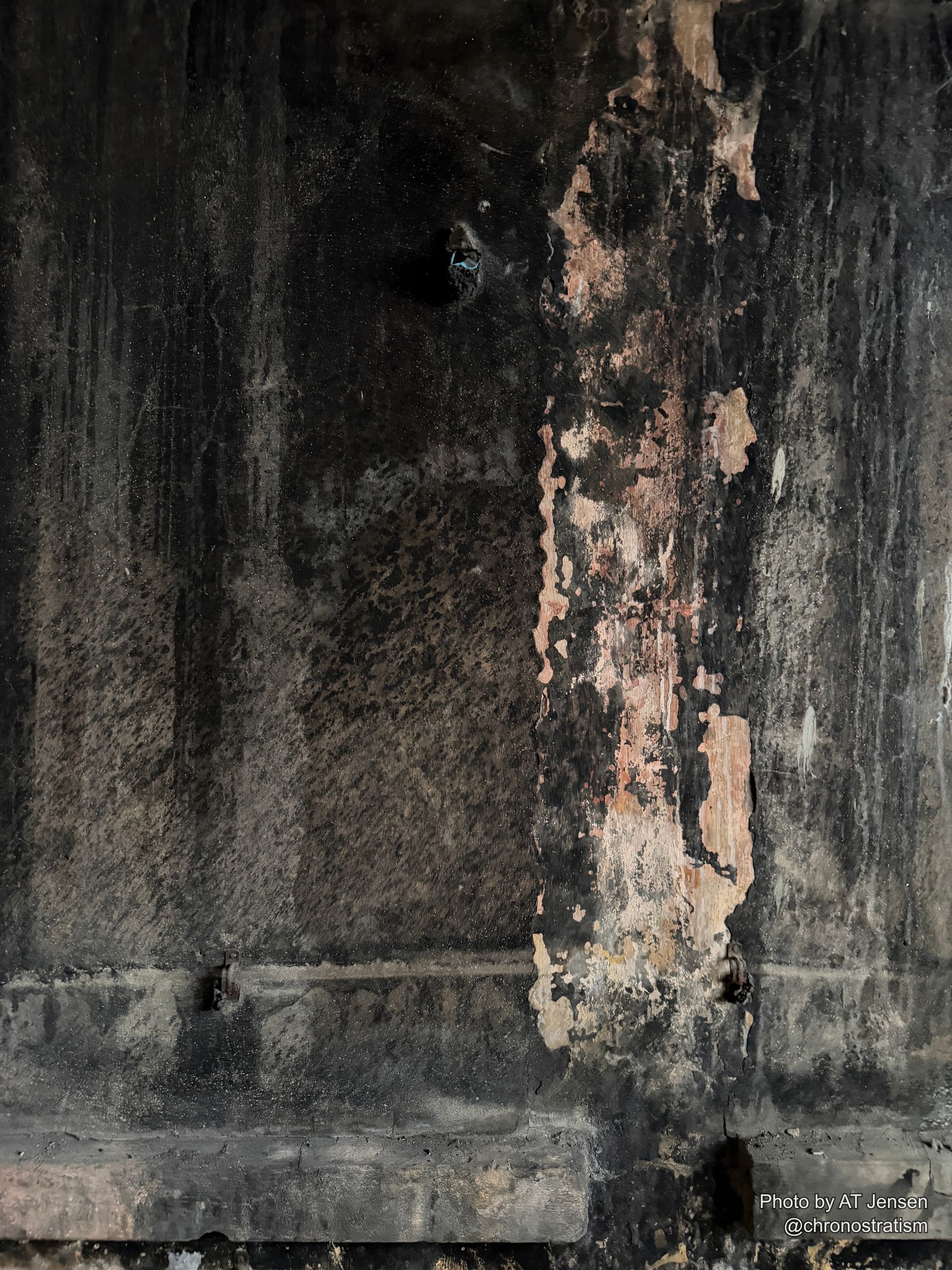 The same soot-darkened wall continues past a rusted hinge, pale plaster breaking through the black crust.