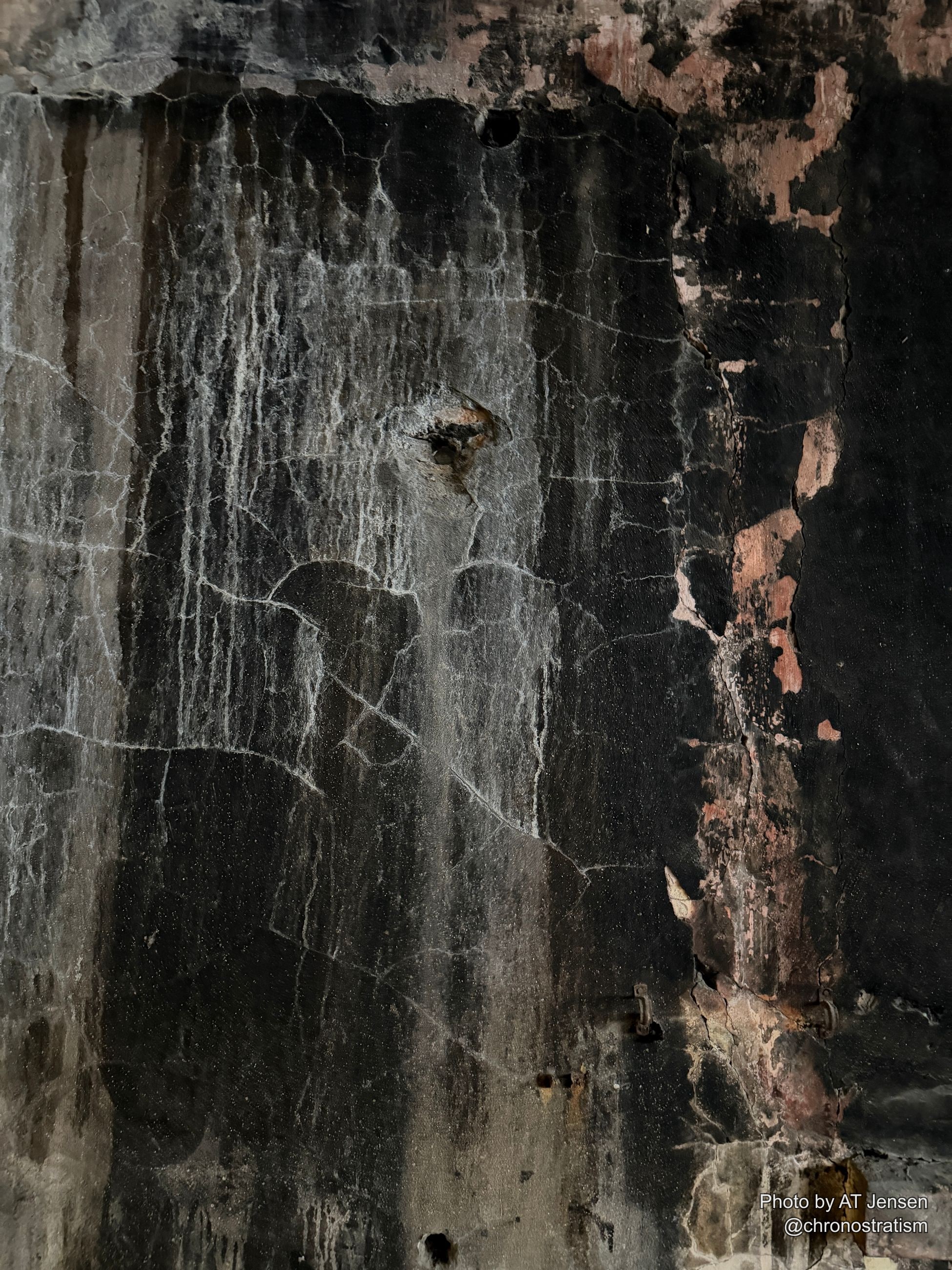 A soot-blackened wall gives way to a raw pink plaster patch breaking through, hairline cracks lacing the surface.
