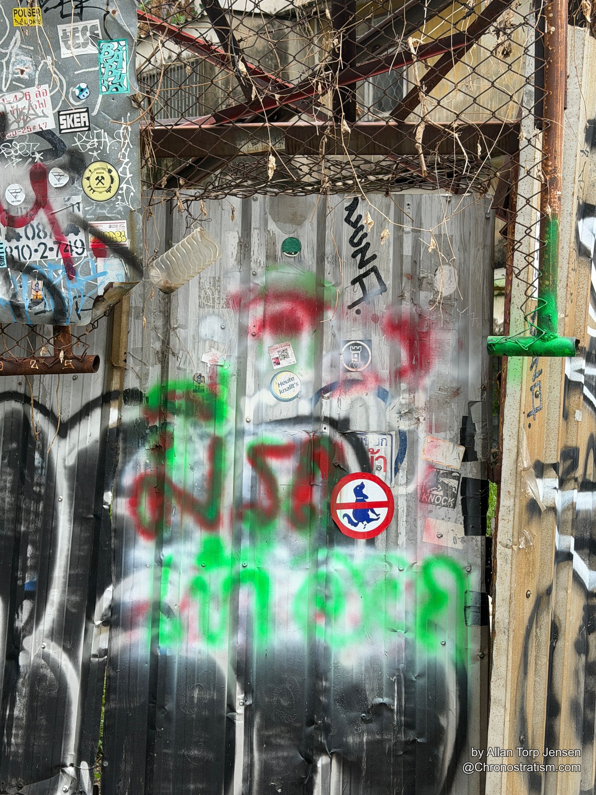 Stickers, spray tags and a hand-painted 'KNOCK' sign crowd a chain-link gate.