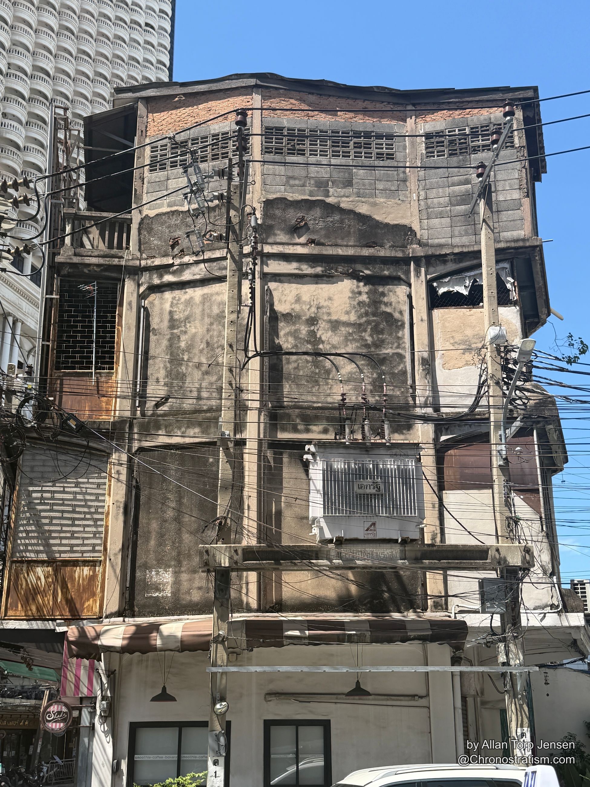 Tangled wires and AC units cover a shophouse facade above a leaning transformer box.