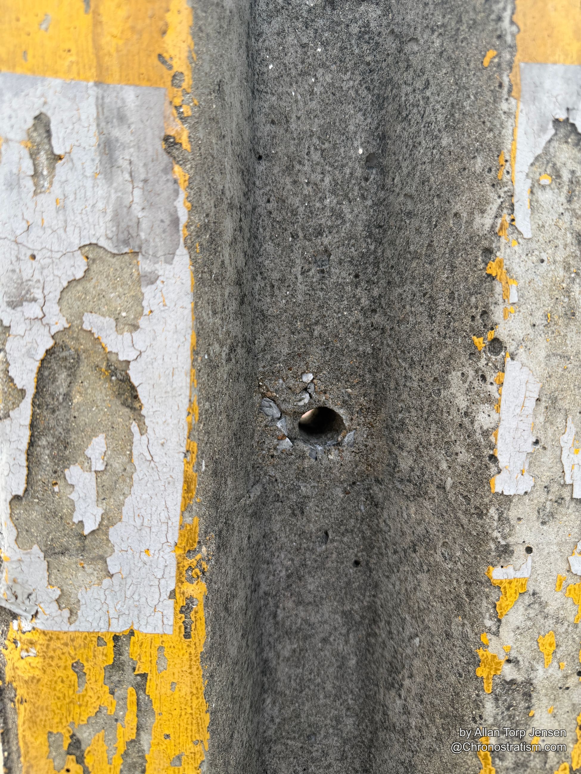 White and yellow paint peels in brittle sheets from a concrete curb, a drilled hole at its center.
