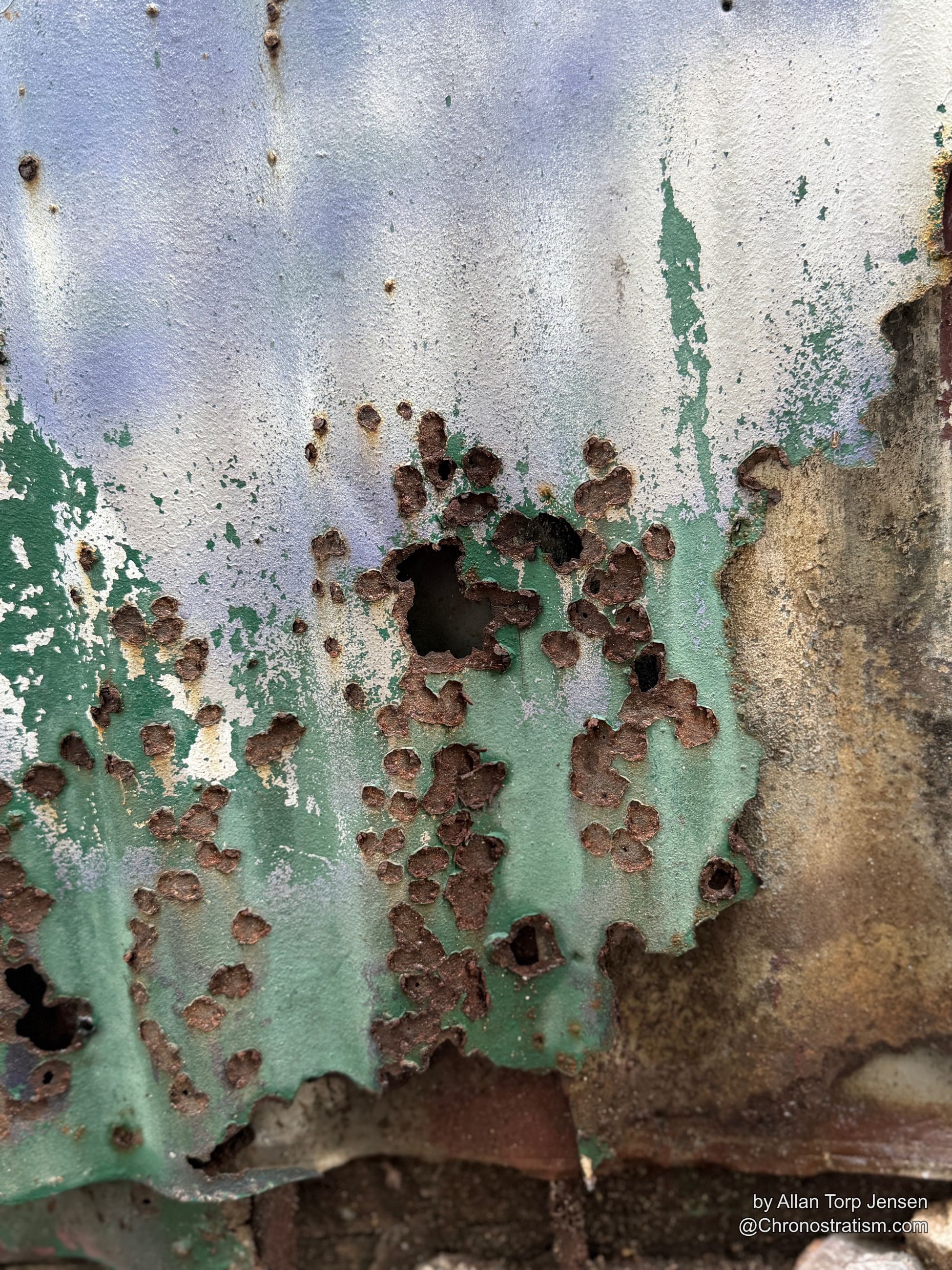 A wide rust-eaten hole breaks through green and lavender paint on corroded metal.