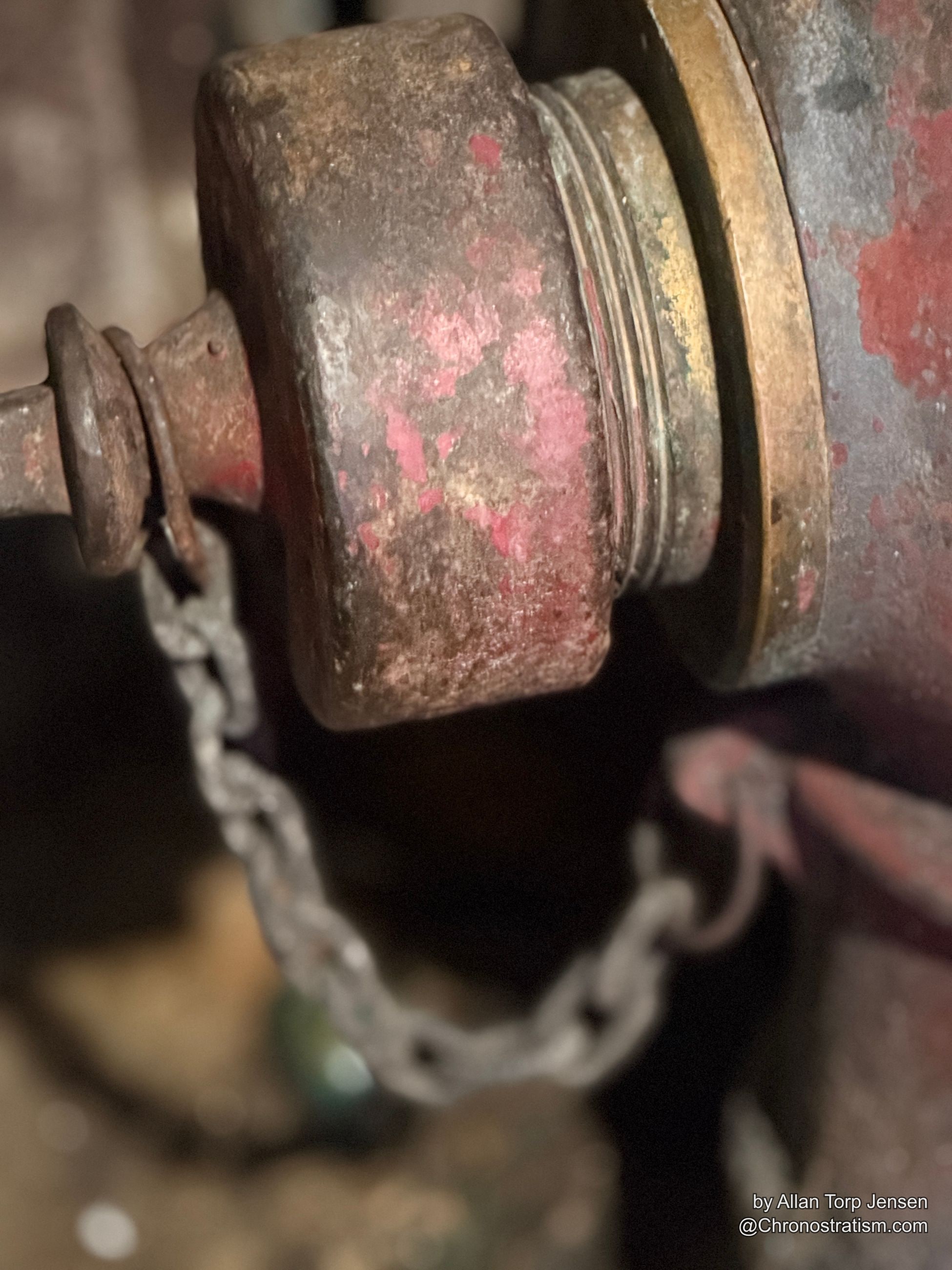 Worn red paint flakes from a brass fire hydrant valve cap eaten by rust.